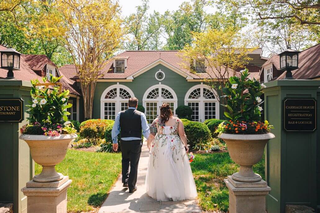 A couple, dressed in wedding attire, walks hand-in-hand along a stone path toward a green house surrounded by manicured gardens and trees.