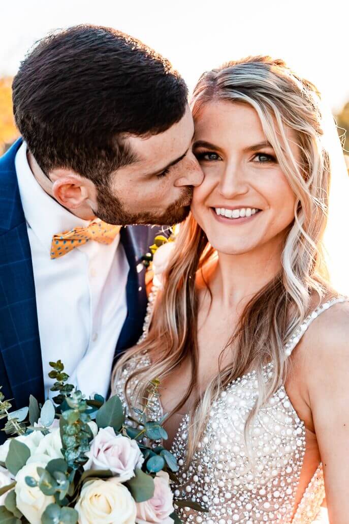 A groom in a blue suit kisses a smiling bride in a beaded dress as she holds a bouquet of roses and greenery.