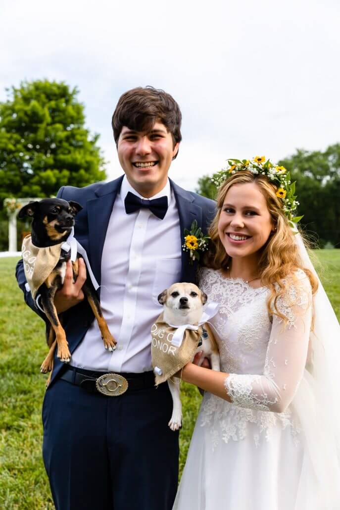 A bride and groom pose outdoors, each holding a small dog wearing a white ribbon labeled 