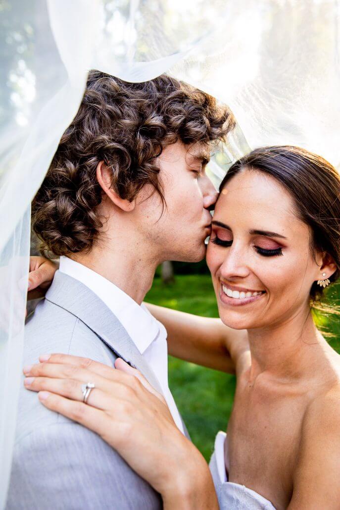A groom in a light gray suit kisses the bride’s forehead as she smiles and holds his shoulder, with her engagement ring visible. They are under a white veil outdoors.
