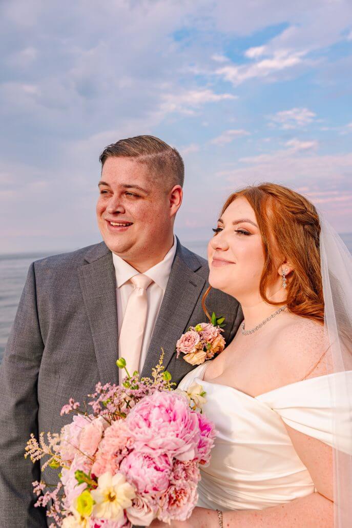 A bride and groom stand together outdoors near water, dressed in wedding attire, holding a bouquet of pink flowers, with a partly cloudy sky in the background.