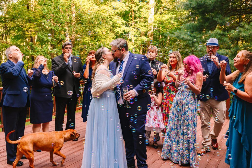 gallery-sophia-0051 A couple shares a kiss on a wooden deck surrounded by guests blowing bubbles; a brown dog is present. The group is dressed formally, and the setting is outdoors with trees in the background.