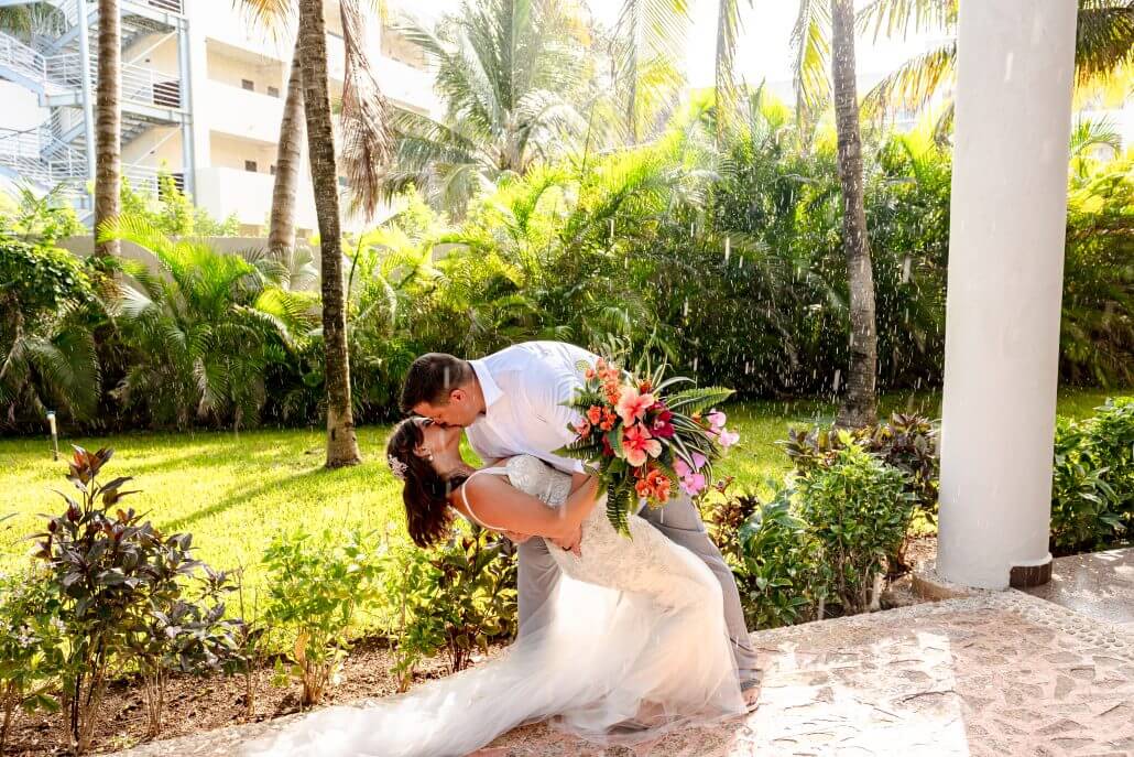 A couple in wedding attire kisses as the groom dips the bride, who holds a colorful bouquet, on a stone patio with palm trees and greenery in the background.