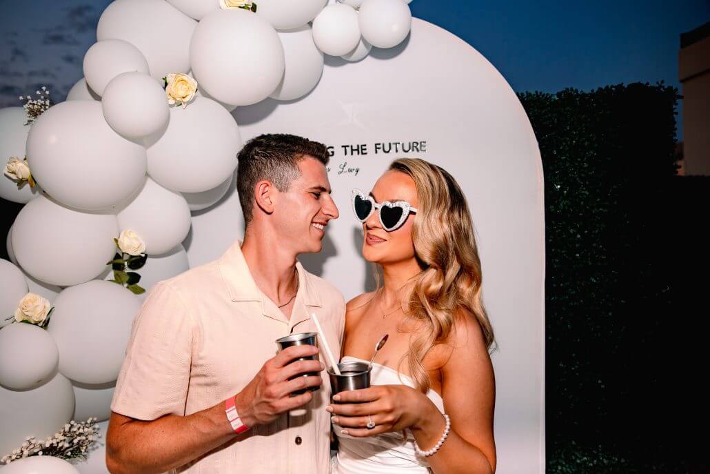 A couple stands in front of a white balloon arch, holding drinks and smiling at each other; the woman wears heart-shaped sunglasses.
