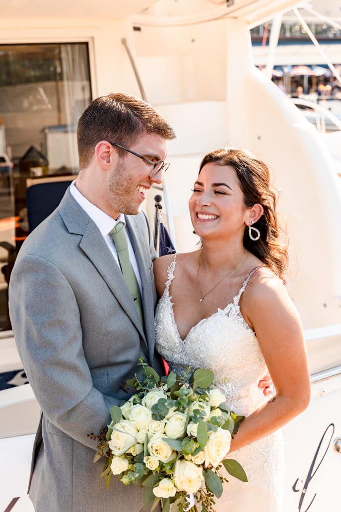 A bride and groom stand close together on a boat, smiling at each other. The bride holds a bouquet of white roses and wears a white lace dress; the groom wears a grey suit and green tie.