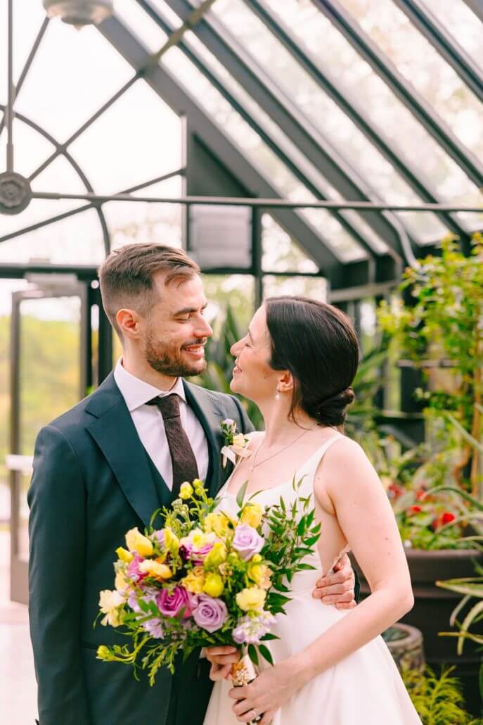 A couple in wedding attire stands indoors, smiling at each other. The bride holds a bouquet of colorful flowers. Green plants and large windows are in the background.