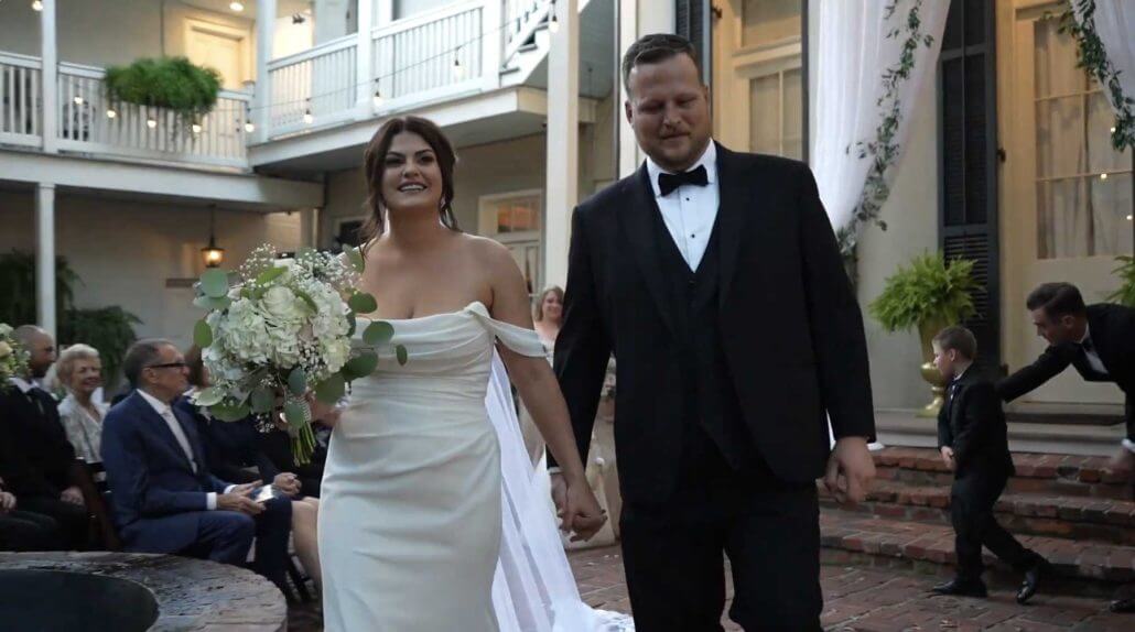 New Orleans wedding photography by Toast: a bride in a white dress and groom in a black tuxedo hold hands and walk down the aisle at an outdoor wedding ceremony, with guests seated and a child in the background.