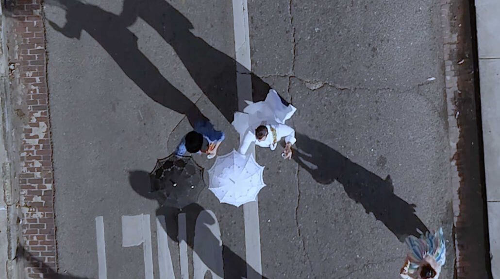 New Orleans wedding photography by Toast: overhead view of two people walking on a street, one holding a white umbrella, casting long shadows; a third person is partially visible at the edge.