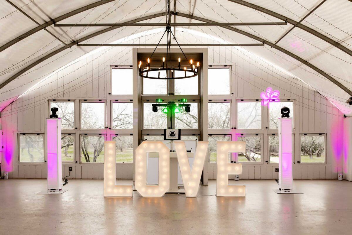 Love-Letters Large illuminated letters spelling "LOVE" stand in the center of a white, modern barn venue with high ceilings, chandeliers, and pink accent lights.