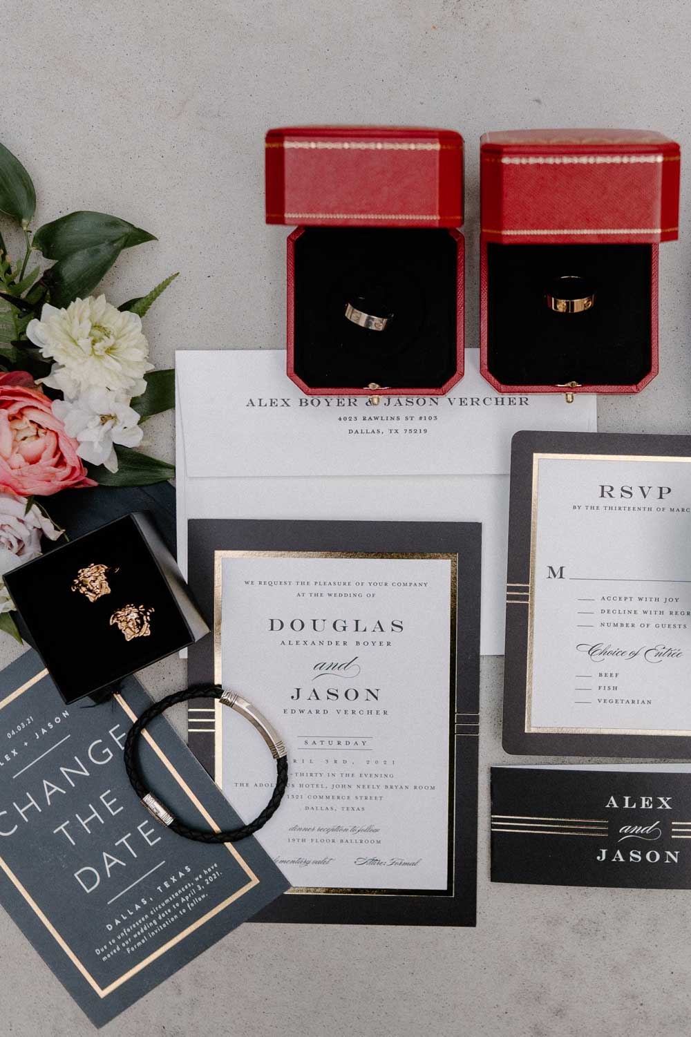 Wedding invitations, RSVP cards, and a change-the-date card arranged with two rings in red boxes, a bracelet in a black box, and flowers on a light surface.