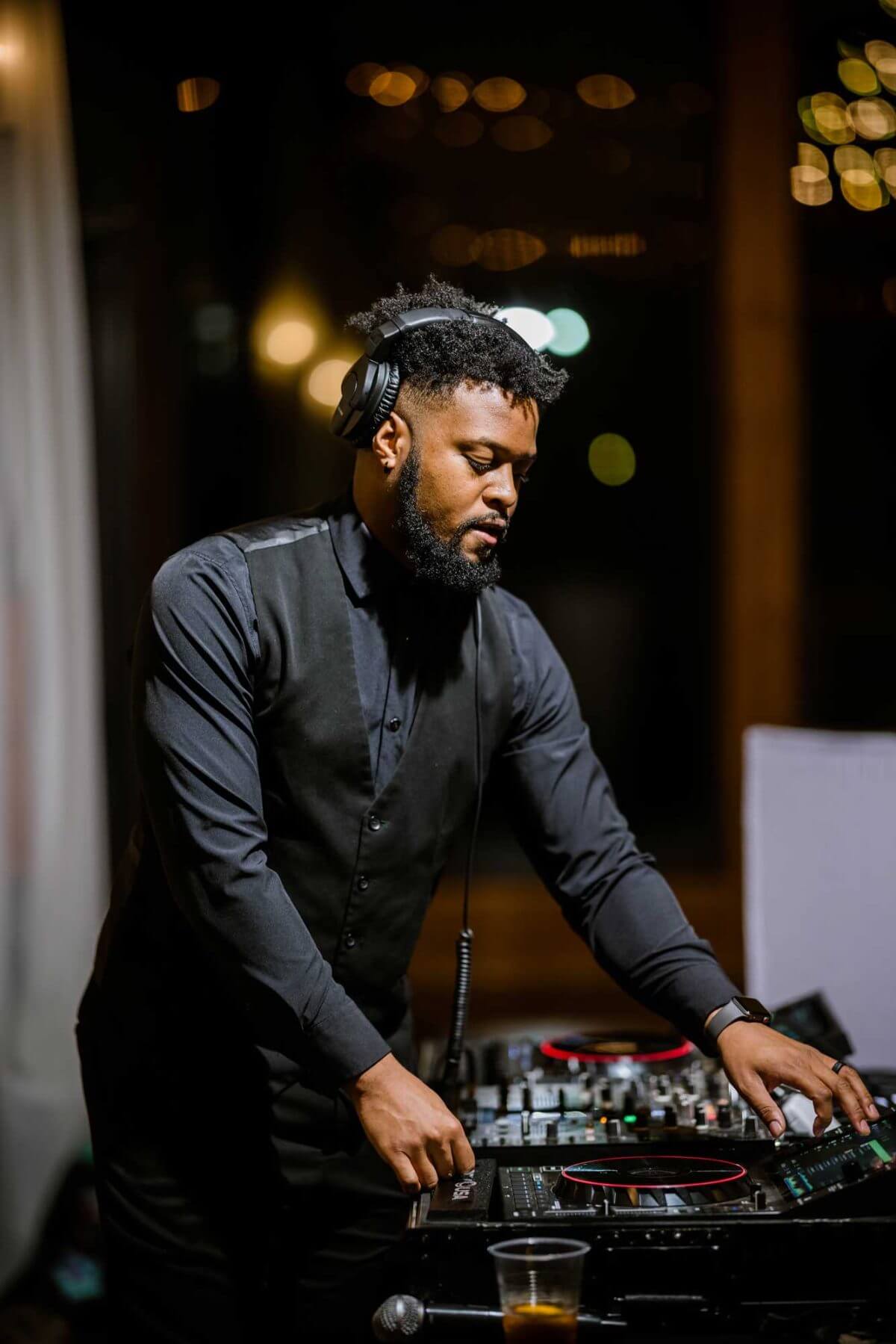 dj-isaiah-in-action A man wearing headphones and a black outfit stands at a DJ mixer, adjusting controls, with a drink on the table in front of him.