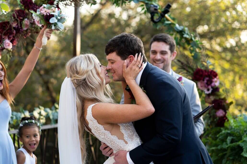 photog-bradt-20201019_KourtneyKerr-183 A bride and groom kiss at their outdoor wedding ceremony, surrounded by smiling guests and floral decorations.