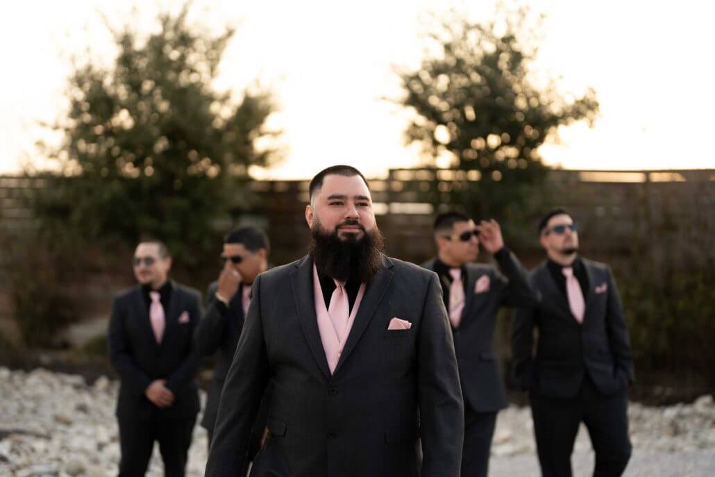 San Antonio wedding photography by Toast: a man in a dark suit with a pink tie and pocket square stands in focus, while four similarly dressed men are blurred in the background outdoors.