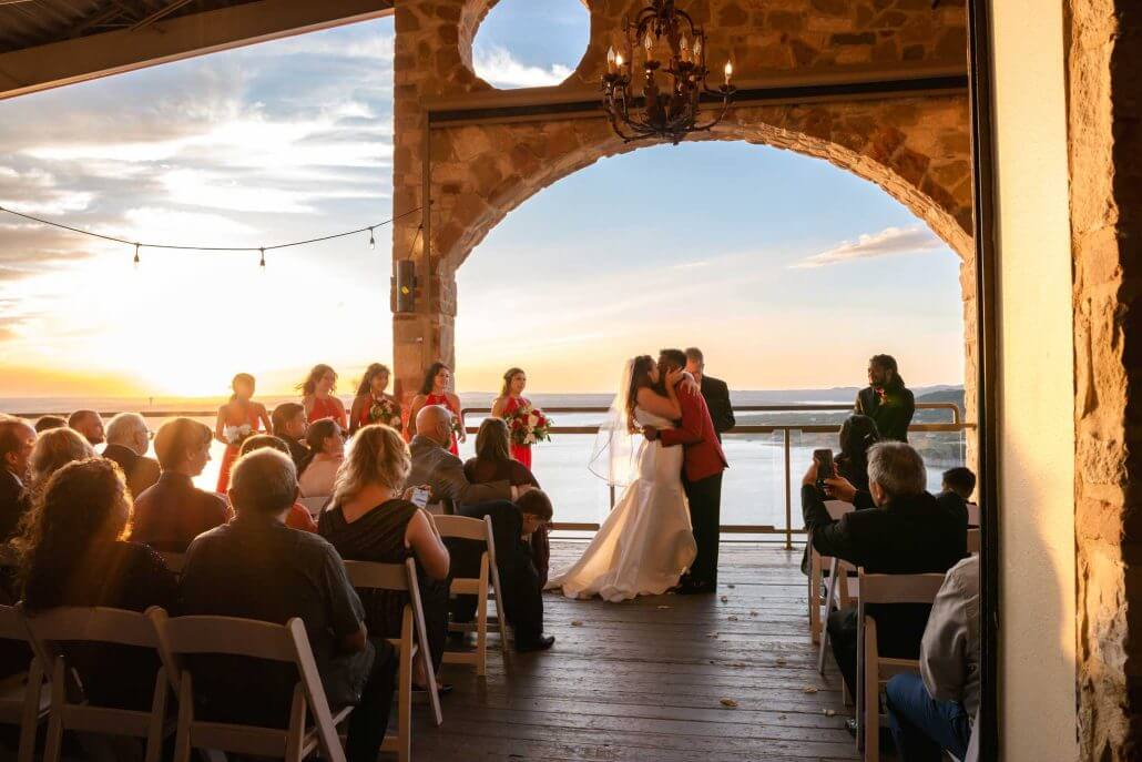 photog-mary_a-Ashlyn-and-Ryan-Ceremony-146 A couple kissing on a deck with people sitting around.