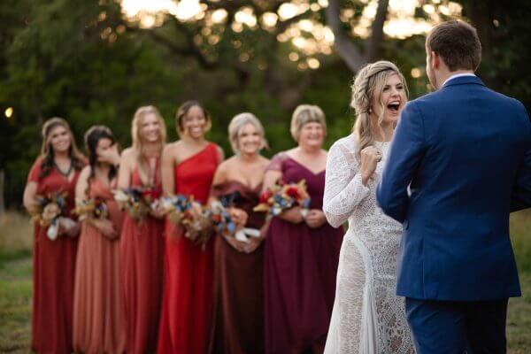 Austin wedding photography by Toast: bride laughs as she sees her groom, with six bridesmaids in red and burgundy behind her.