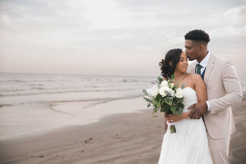 South Texas wedding photography by Toast: a bride and groom stand on a beach; the groom hugs and kisses the bride on the head as she holds a bouquet of white flowers.