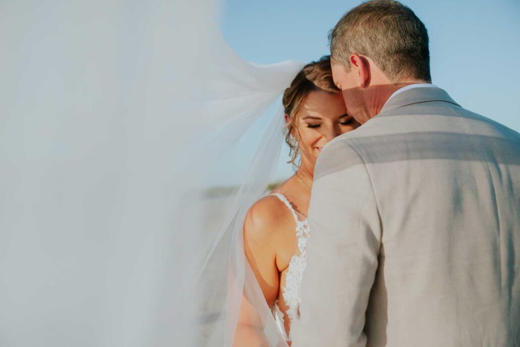 South Texas wedding photography by Toast: a bride and groom stand close together outdoors, the bride smiling with her eyes closed, partially covered by a flowing veil, both dressed in formal attire.