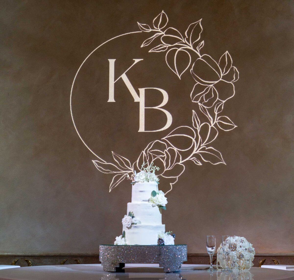 A monogram with a floral design projected on the wall behind a tiered wedding cake.