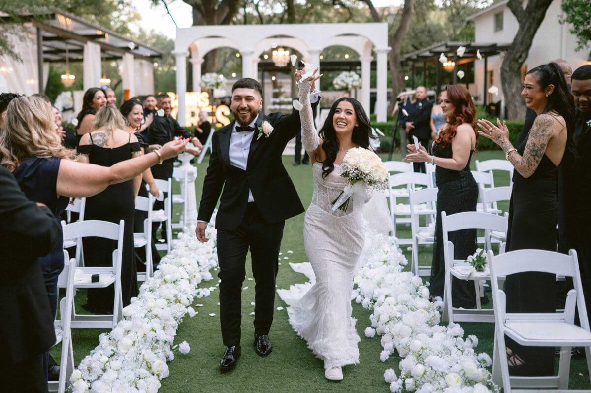 wedding-ceremony A bride and groom walk down an outdoor aisle lined with white flowers as guests on both sides applaud and celebrate their wedding.