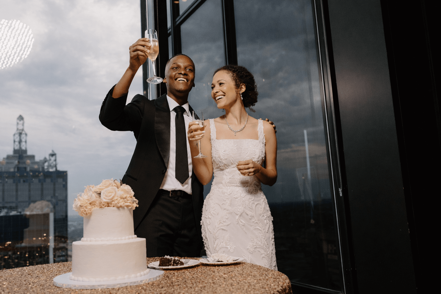 A bride and groom in formal attire stand beside a decorated wedding cake, smiling and holding up glasses for a toast by a large window with a city view.