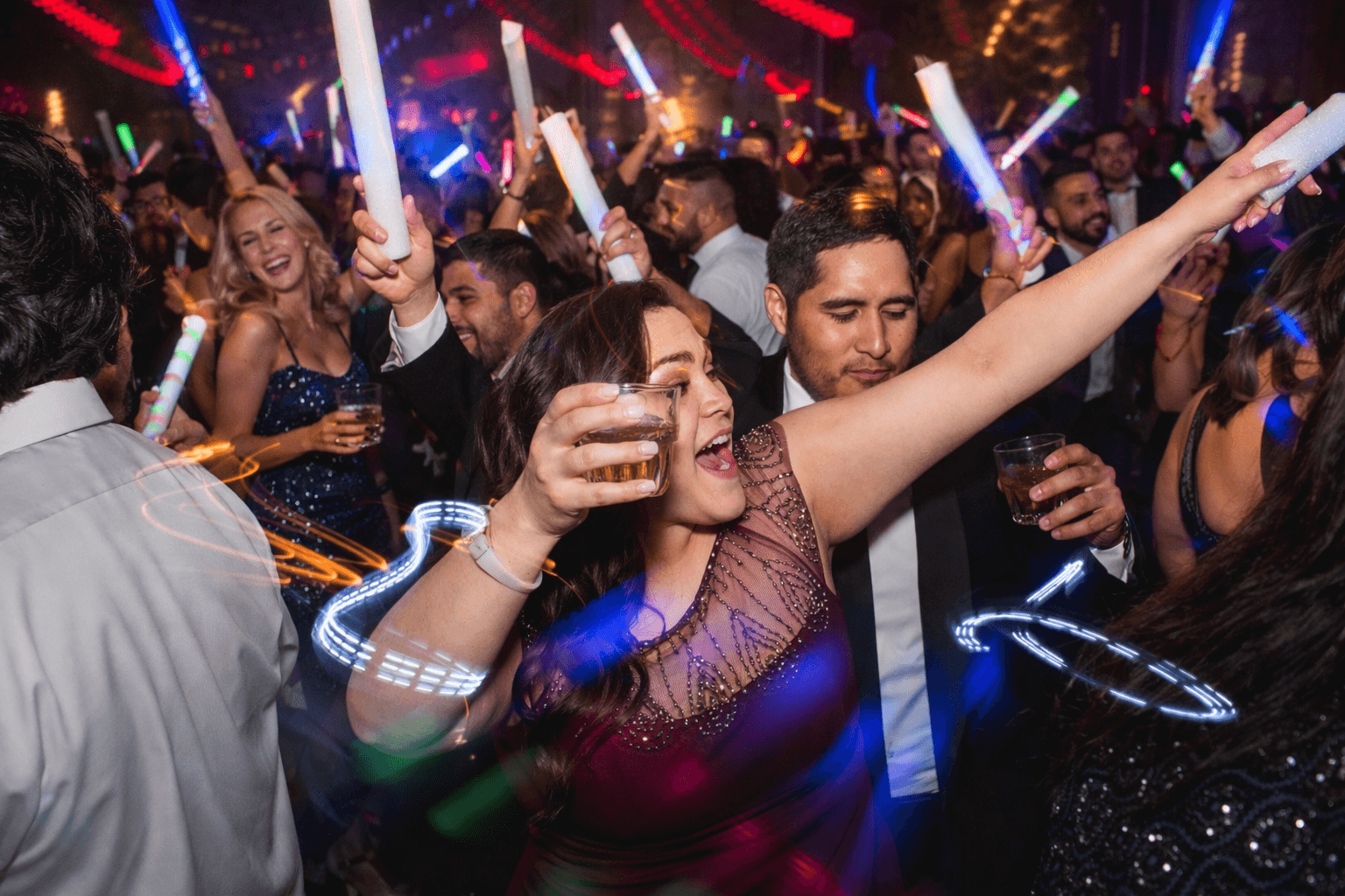 Colorful nightclub scene with a woman in a sequined dress dancing and raising her arms, surrounded by guests and glowing light sticks in a lively party atmosphere.