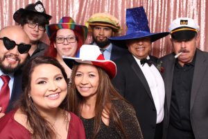 Family in Photo Booth Pink Backdrop Eight people posing in front of a pink sequin backdrop, wearing formal attire and various novelty hats, including a wizard hat, rainbow hat, and captain’s hat with a fake cigar.