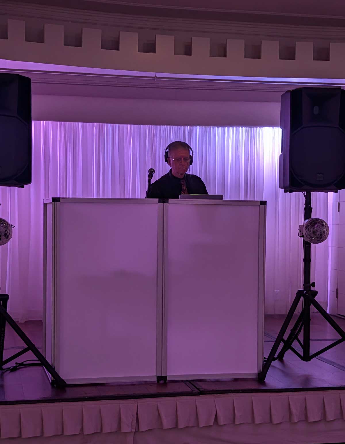Dennis Falcone behind a white DJ booth with purple lighting at a Long Island event, wearing headphones and a dark shirt, surrounded by speakers.