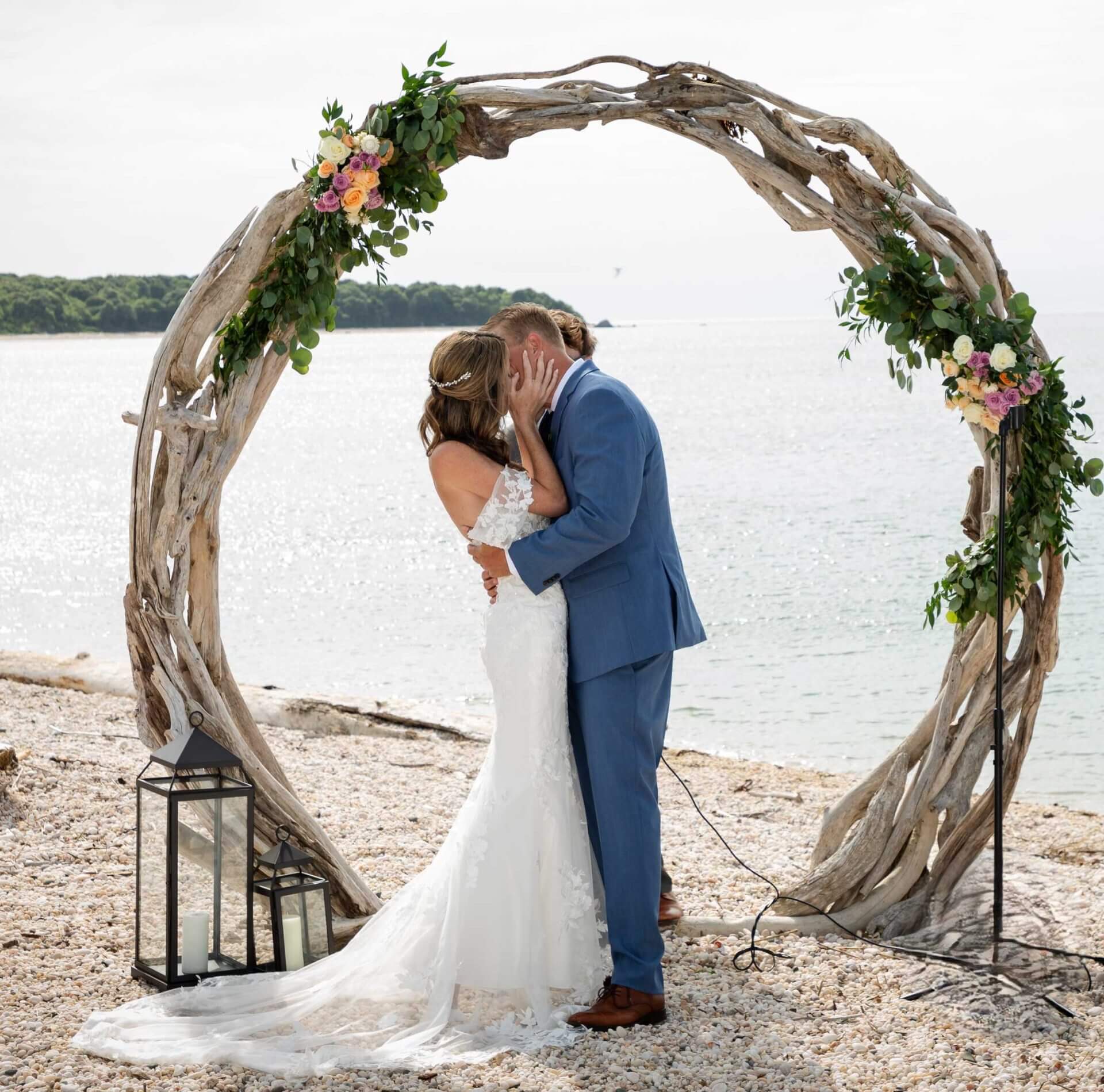 A couple in wedding attire kisses beneath a circular wooden arch decorated with flowers and greenery on a beach, with the ocean in the background.