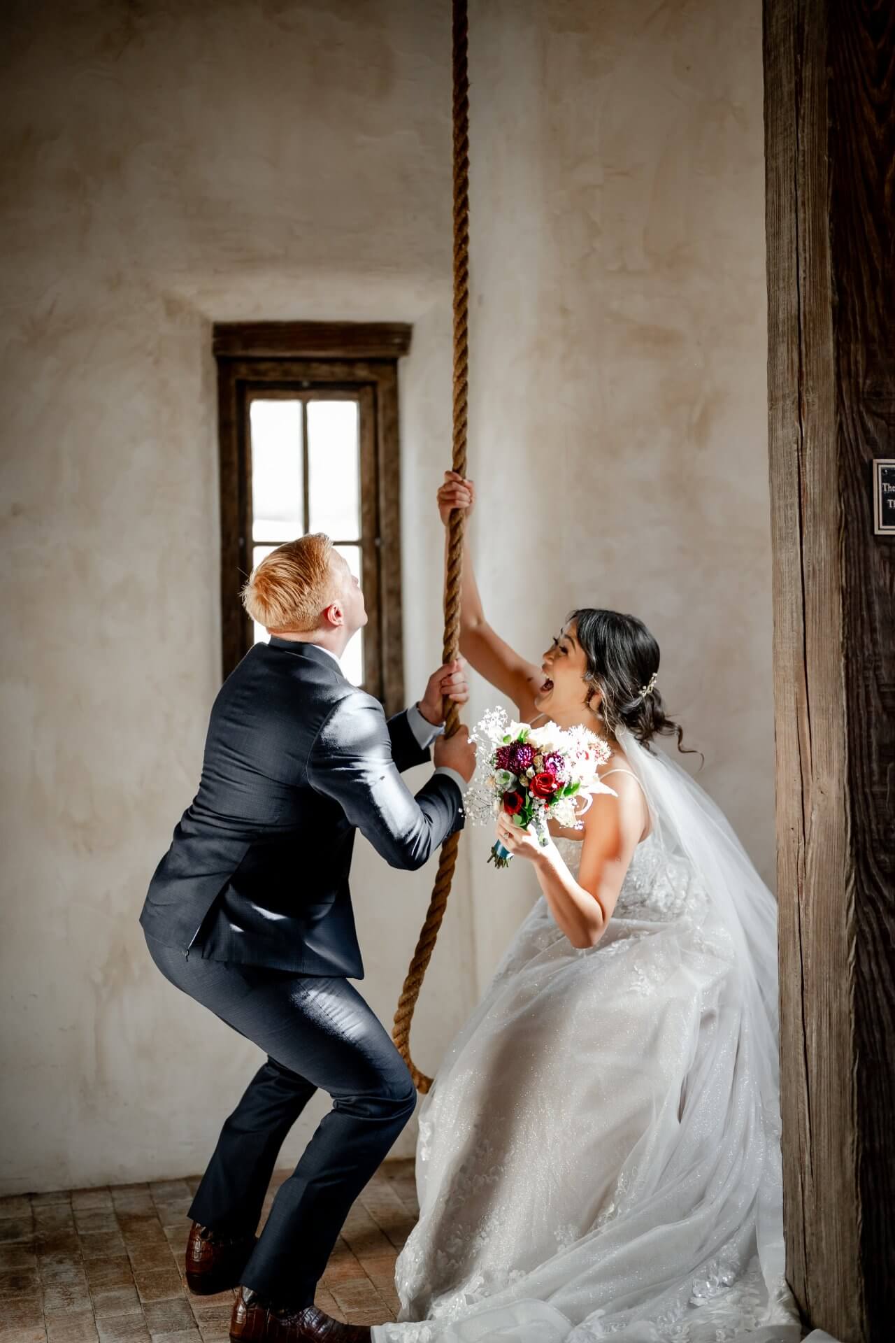 A7400685 A bride in a white dress holding a bouquet and a groom in a suit pull a large rope together indoors, with sunlight streaming through a nearby window.
