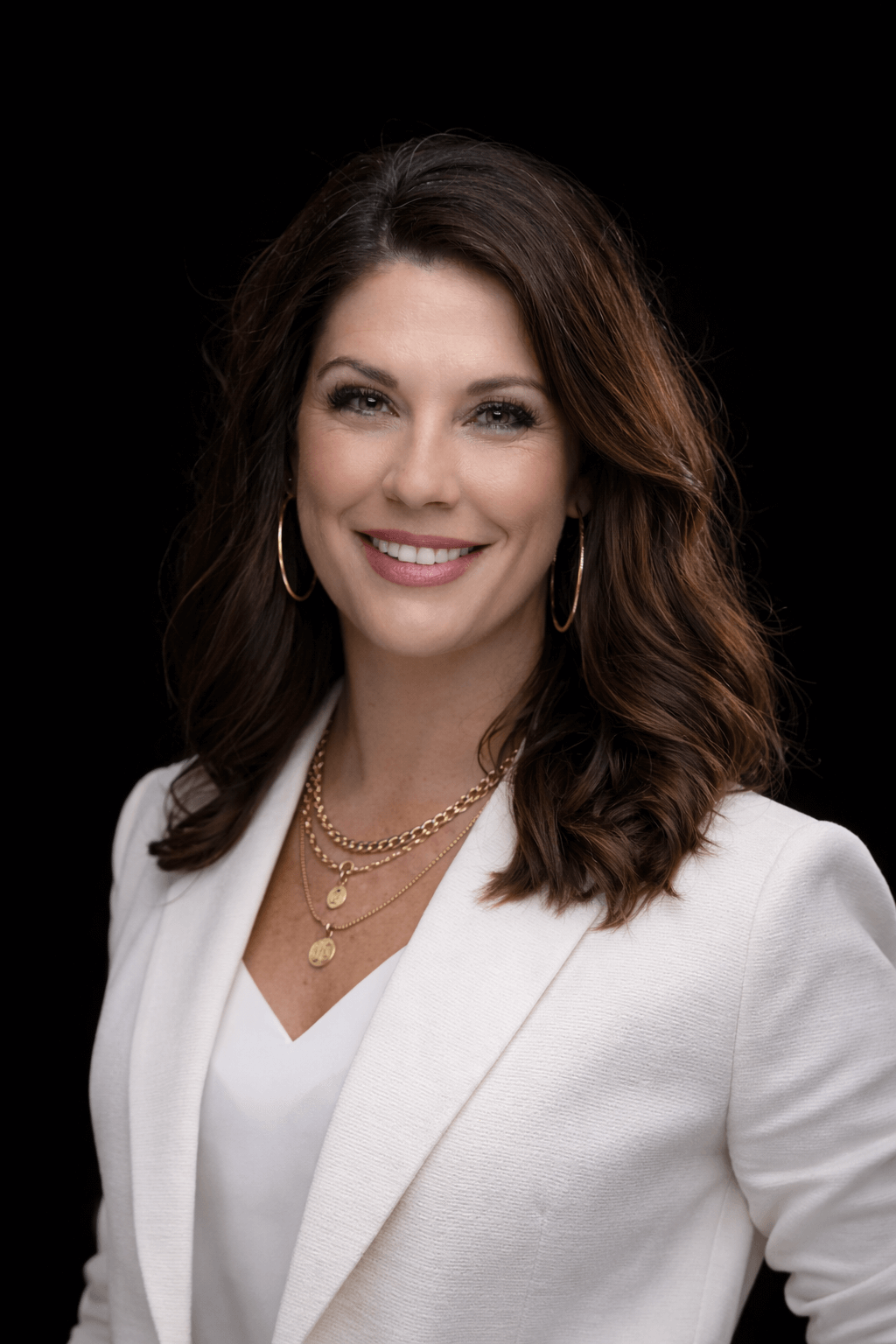 Woman with long brown hair wearing a white blazer, gold hoop earrings, and layered gold necklaces, smiling in front of a black background.