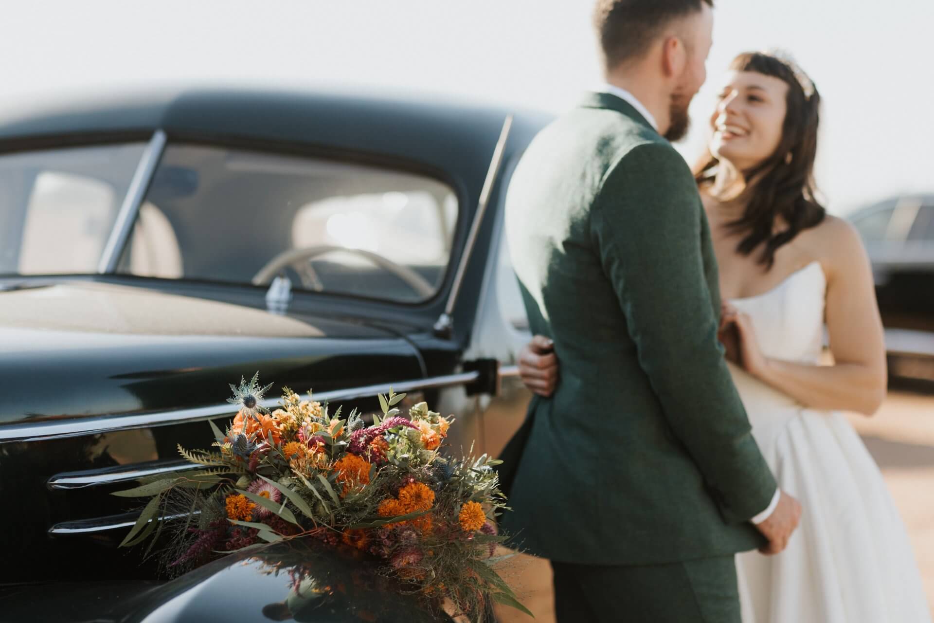 Austin-wedding-photography-ceremony-bride-groom-portaits A bride and groom stand smiling beside a vintage black car with a colorful bouquet placed on the car's hood.