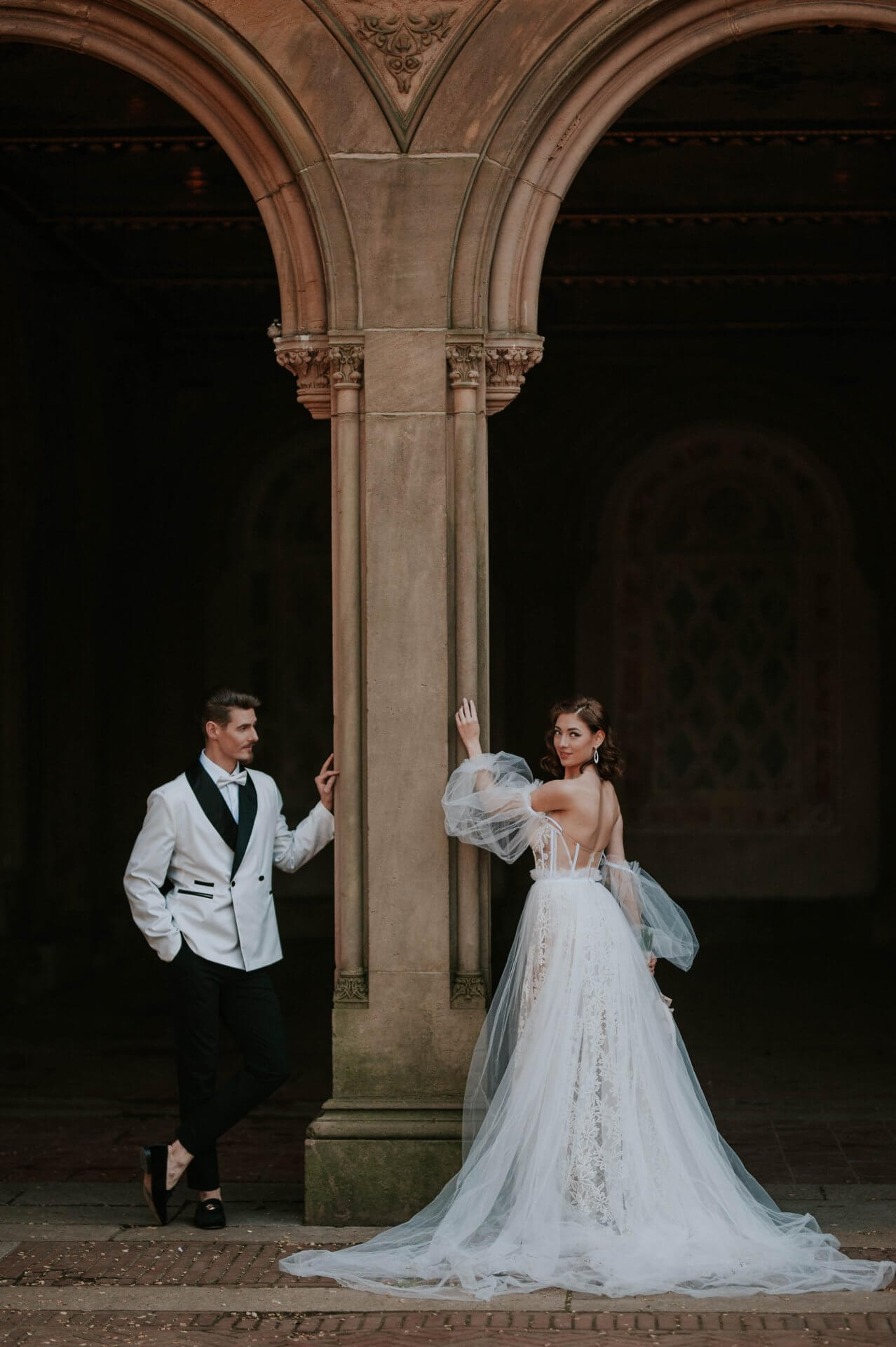 Long-island-wedding-bride-groom-portraits-photography A man in a white tuxedo jacket and a woman in a white wedding gown pose on opposite sides of a stone archway in an elegant, dimly lit setting.