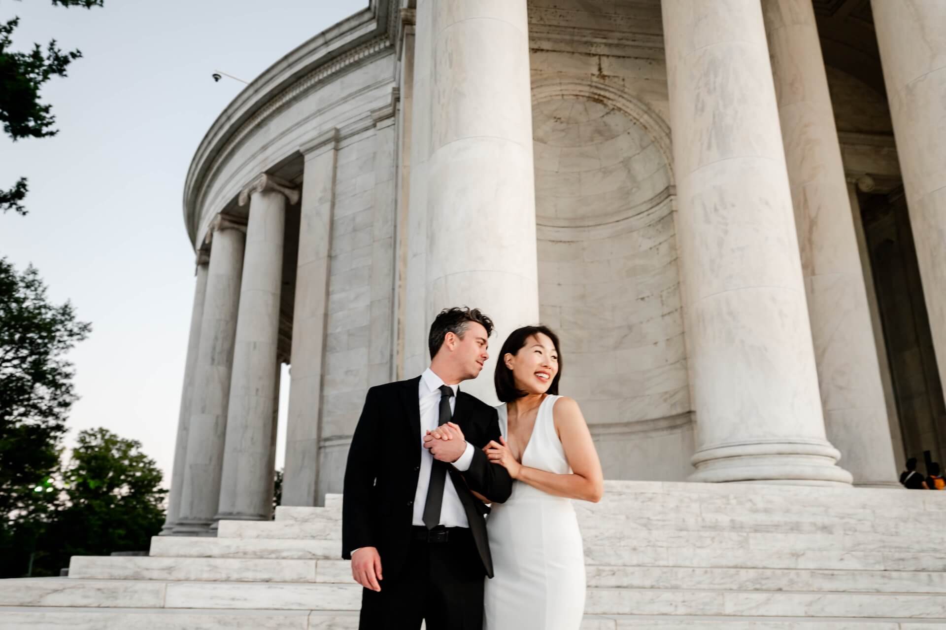 Washington-photography-bride-groom-portraits-memorial A man in a black suit and a woman in a white dress stand together on marble steps in front of large columns, both smiling and holding hands.