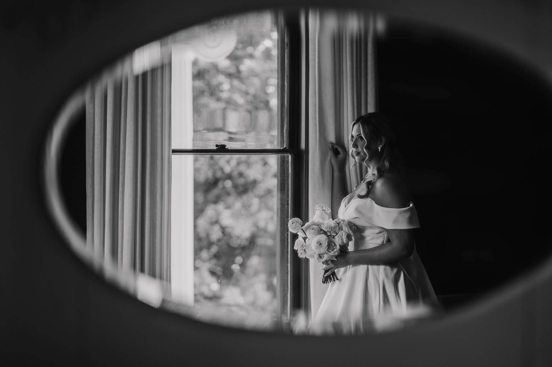 ausin-bride-getting Ready-photography A bride in an off-the-shoulder dress holds a bouquet and stands by a window, seen through an oval mirror in black and white.