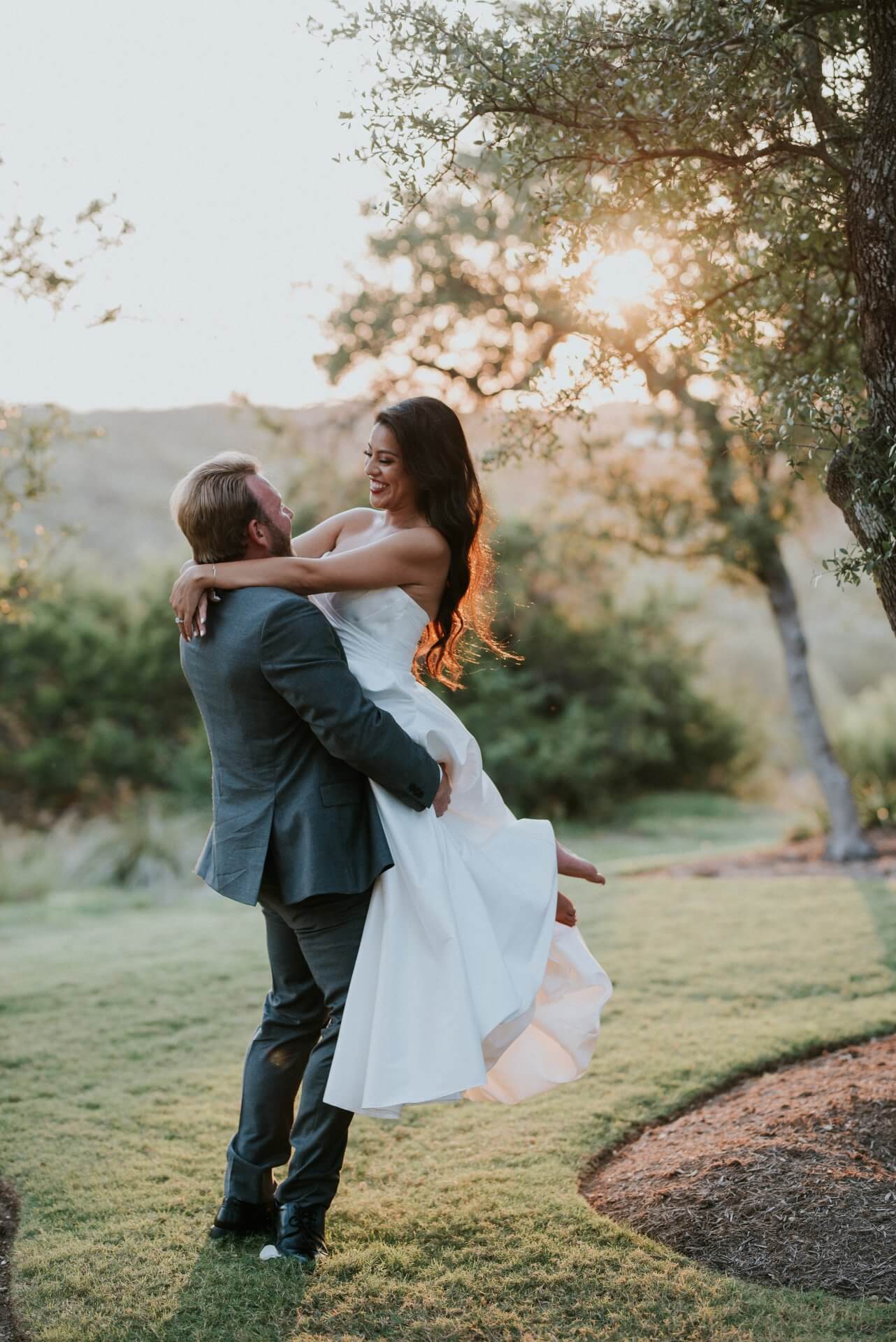 austin-wedding-couple-portraits-bride-groom A groom in a suit lifts his bride in a white dress outdoors at sunset, both smiling, with trees and greenery in the background.