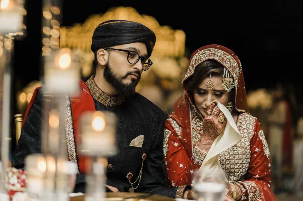 A bride and groom in traditional South Asian wedding attire sit together; the bride holds a cloth to her face while the groom looks on. Candles are blurred in the foreground.
