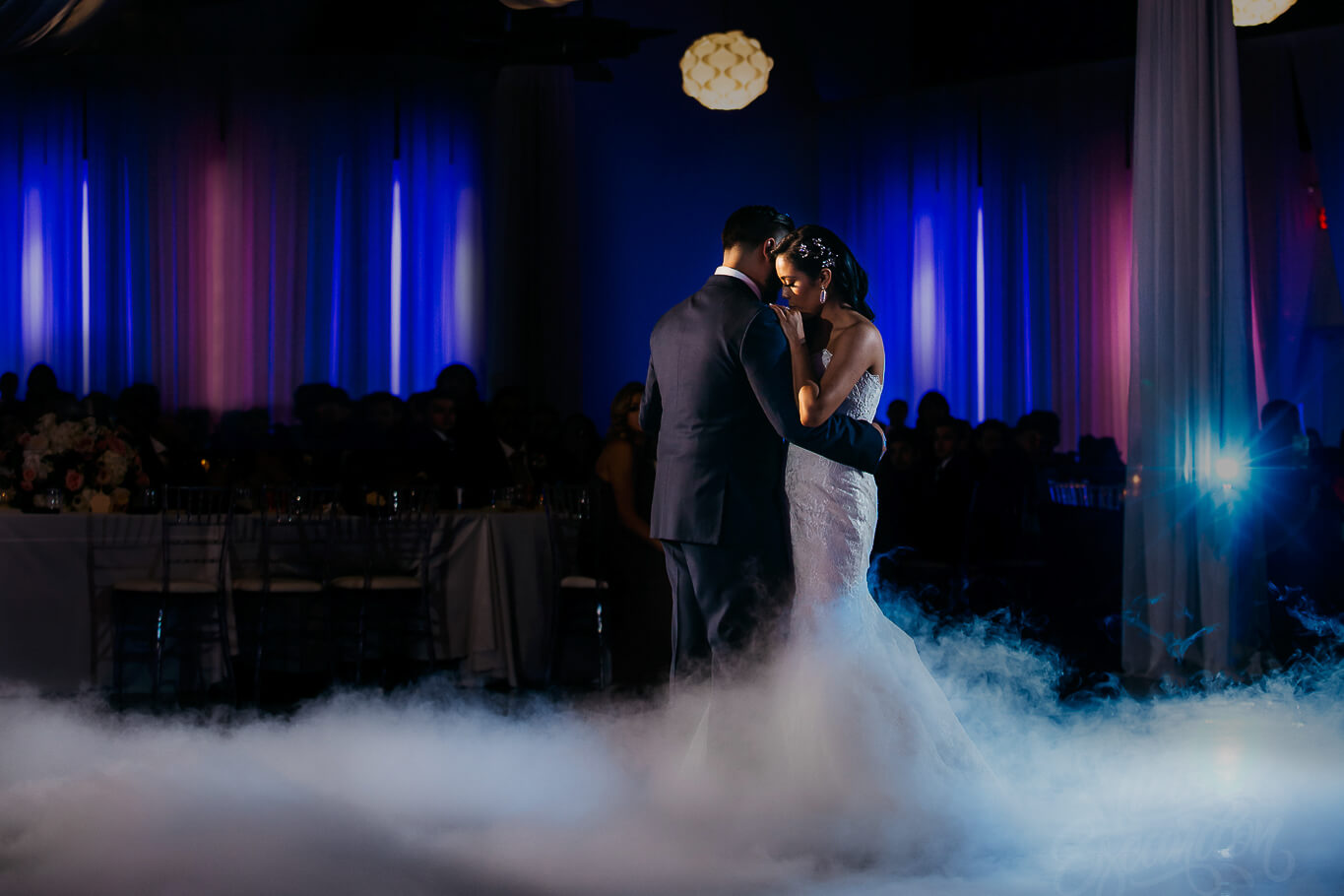 A bride and groom share a first dance on a fog-covered floor in a dimly lit venue with purple and blue lighting.
