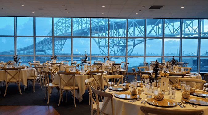 Round banquet tables with place settings are arranged for a wedding reception in a dining room with large windows, overlooking a bridge and water on a cloudy day.