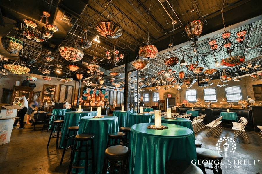 Spacious event venue perfect for a wedding reception, featuring teal-covered tables, black stools, and numerous stained glass lamps hanging from the ceiling, all illuminated by warm ambient indoor lighting.