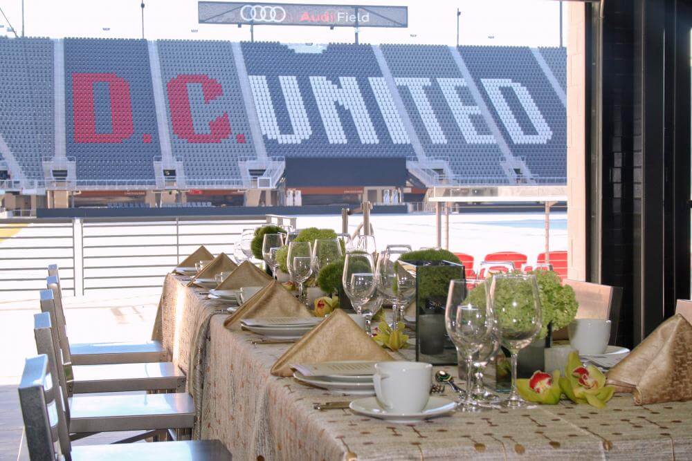A table set for a formal meal, perfect for a wedding reception, overlooks the seating area of Audi Field stadium, with "D.C. United" displayed on the stands in red and white.
