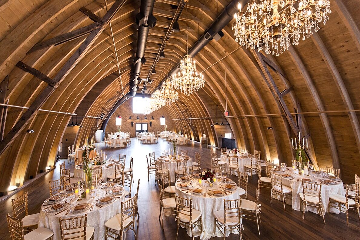 A spacious, arched wooden banquet hall set for a wedding reception, featuring round tables, gold chairs, sparkling chandeliers, and elegant floral centerpieces.