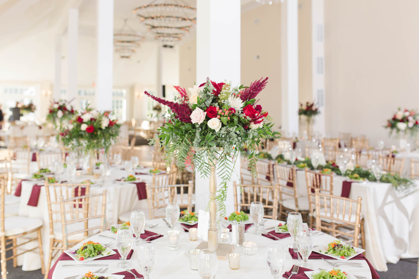 A round table set for a wedding reception with gold chairs, white tablecloth, burgundy napkins, glassware, and a tall floral centerpiece featuring red, white, and pink flowers with greenery.