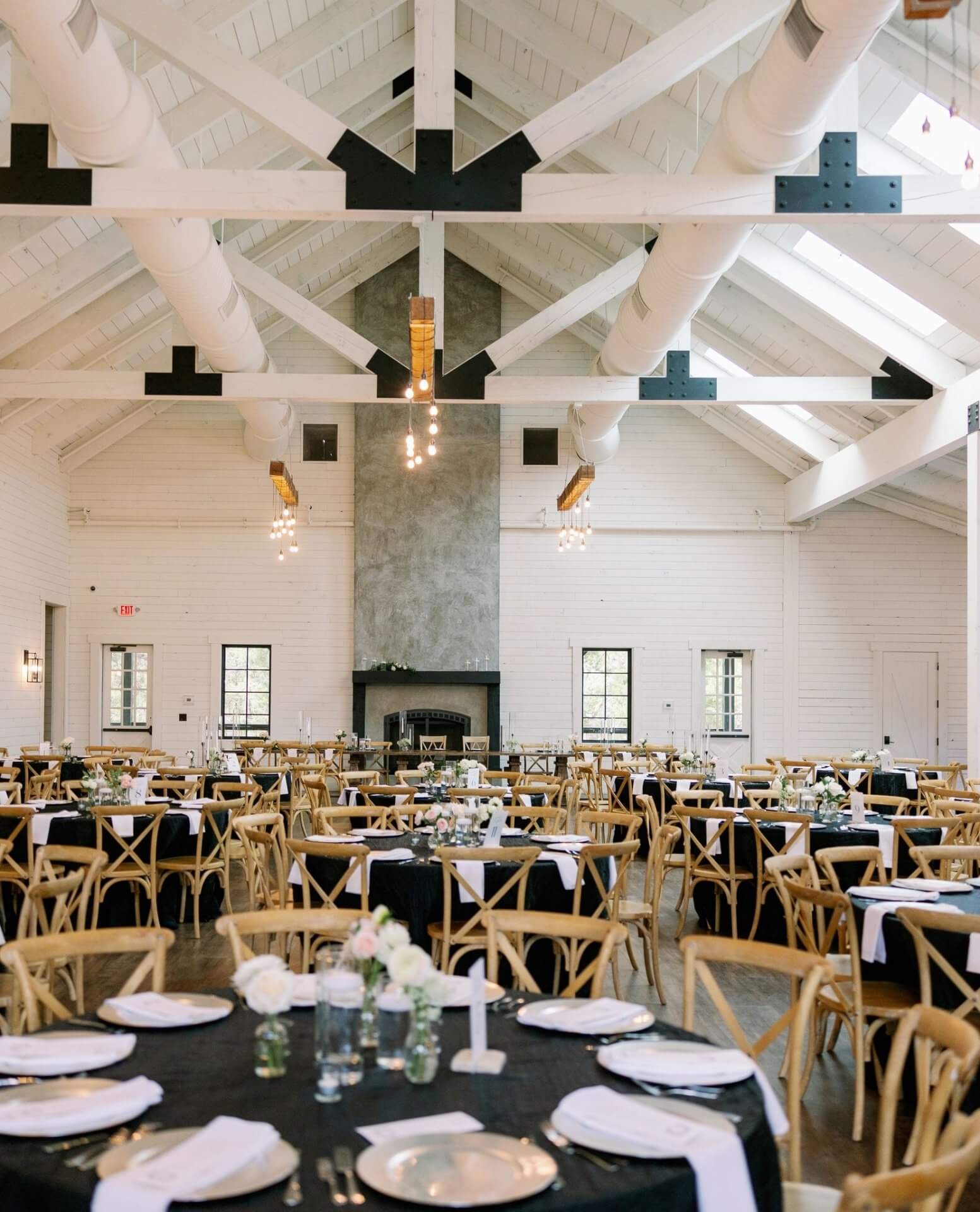 A spacious event hall with high vaulted ceilings, wooden beams, and set tables—perfect for a wedding reception—adorned with black tablecloths, white plates, and simple floral centerpieces.