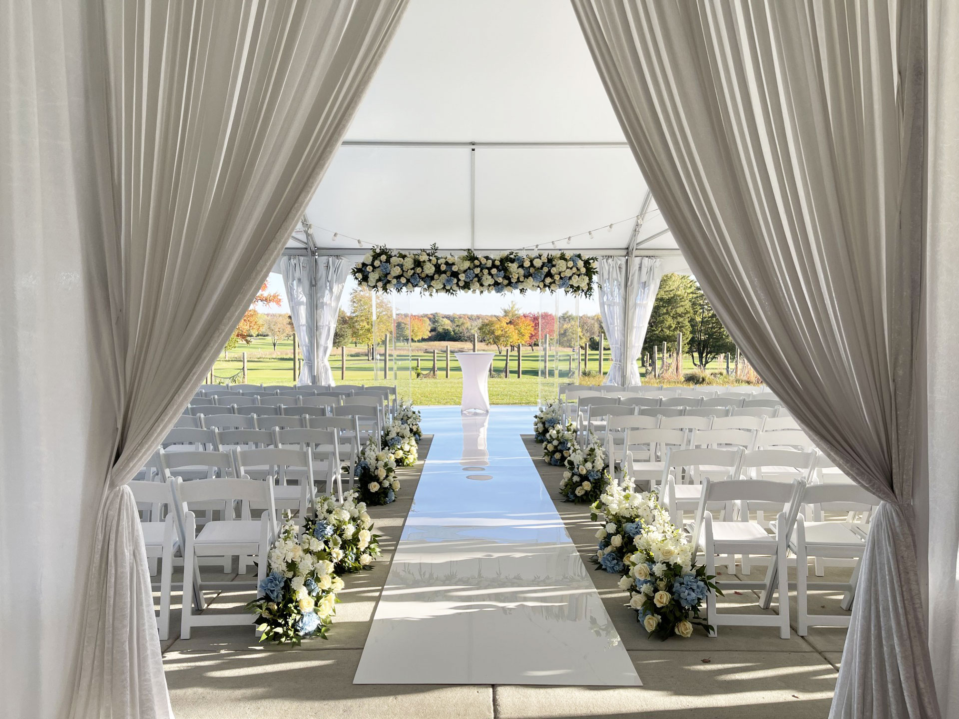 A white tented wedding ceremony setup with rows of white chairs, flower arrangements along the aisle, and an archway decorated with white flowers, perfect for transitioning into a romantic wedding reception.