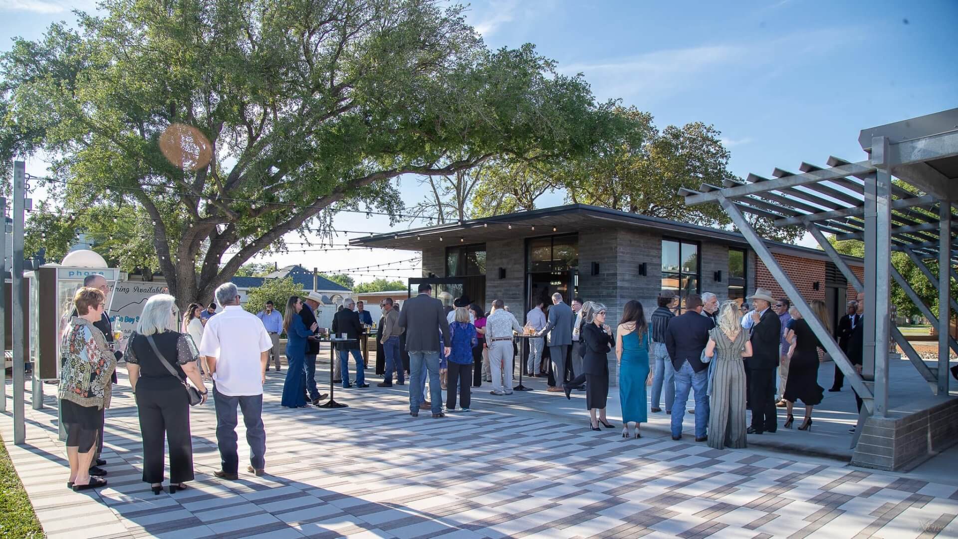 A group of people stands in line and gathers outside a modern building with large windows on a sunny day, surrounded by trees and a tiled courtyard, waiting to enter a wedding reception.