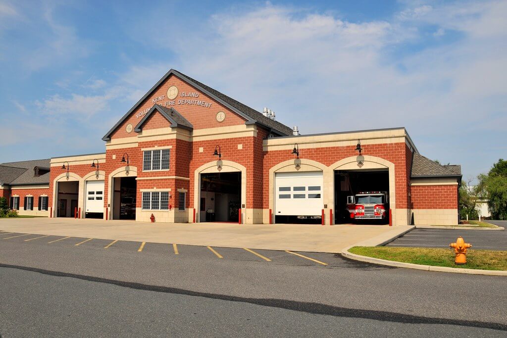 A brick fire station with four garage bays, two open to reveal fire trucks inside. The building, labeled “Fire Department,” sits before parking spaces and a fire hydrant—an unconventional spot for a memorable wedding reception.