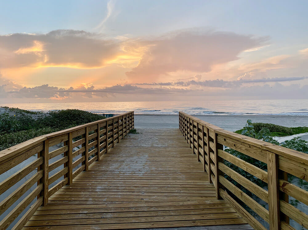 A wooden boardwalk leads to a sandy beach, perfect for a wedding reception, with gentle waves under a colorful sunset sky and scattered clouds.