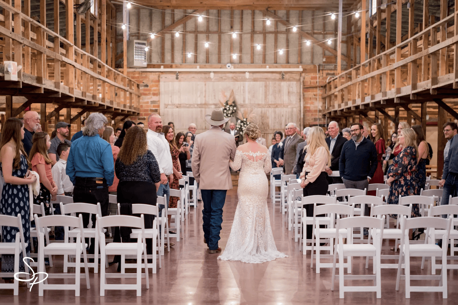 A bride and groom walk down the aisle together in a rustic barn venue, decorated with string lights and ready for a charming wedding reception, as guests stand on either side.