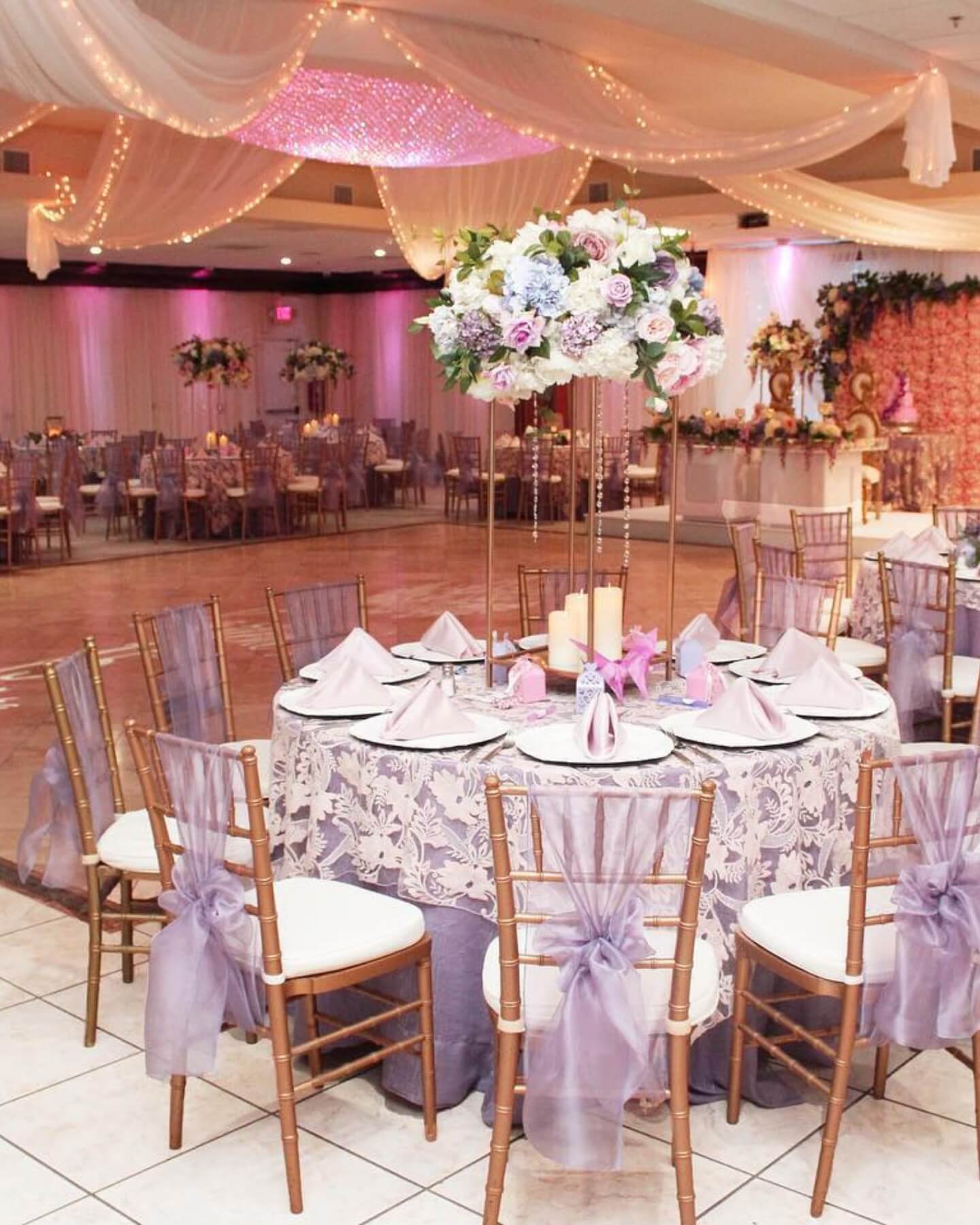 A banquet hall decorated for a wedding reception with round tables, lavender chair sashes, floral centerpieces, draped ceilings, and string lights.