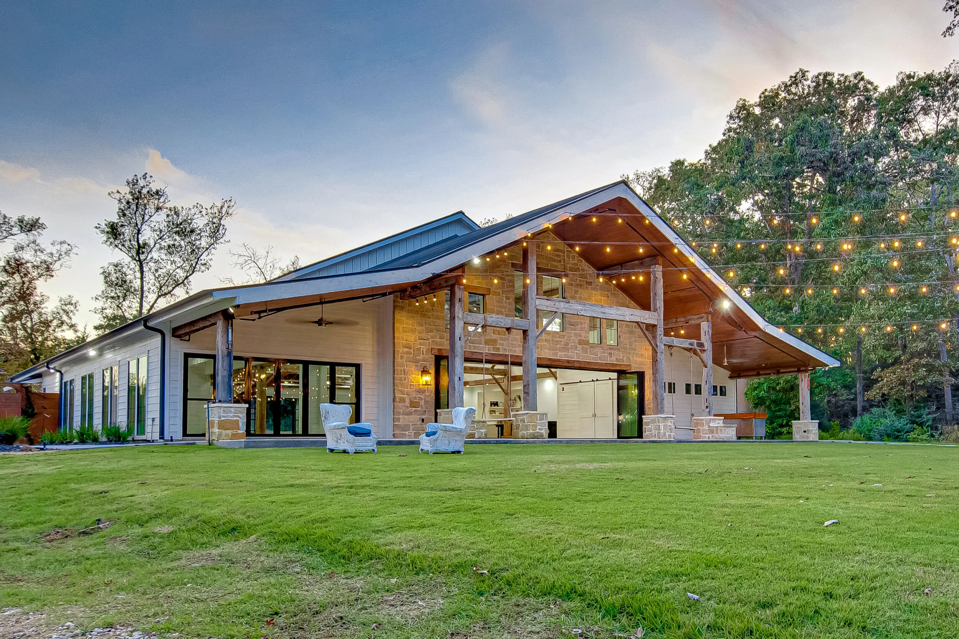 Modern barn-style building with large glass doors, outdoor string lights, and patio seating—perfect for a wedding reception—set on a grassy lawn with trees in the background at dusk.